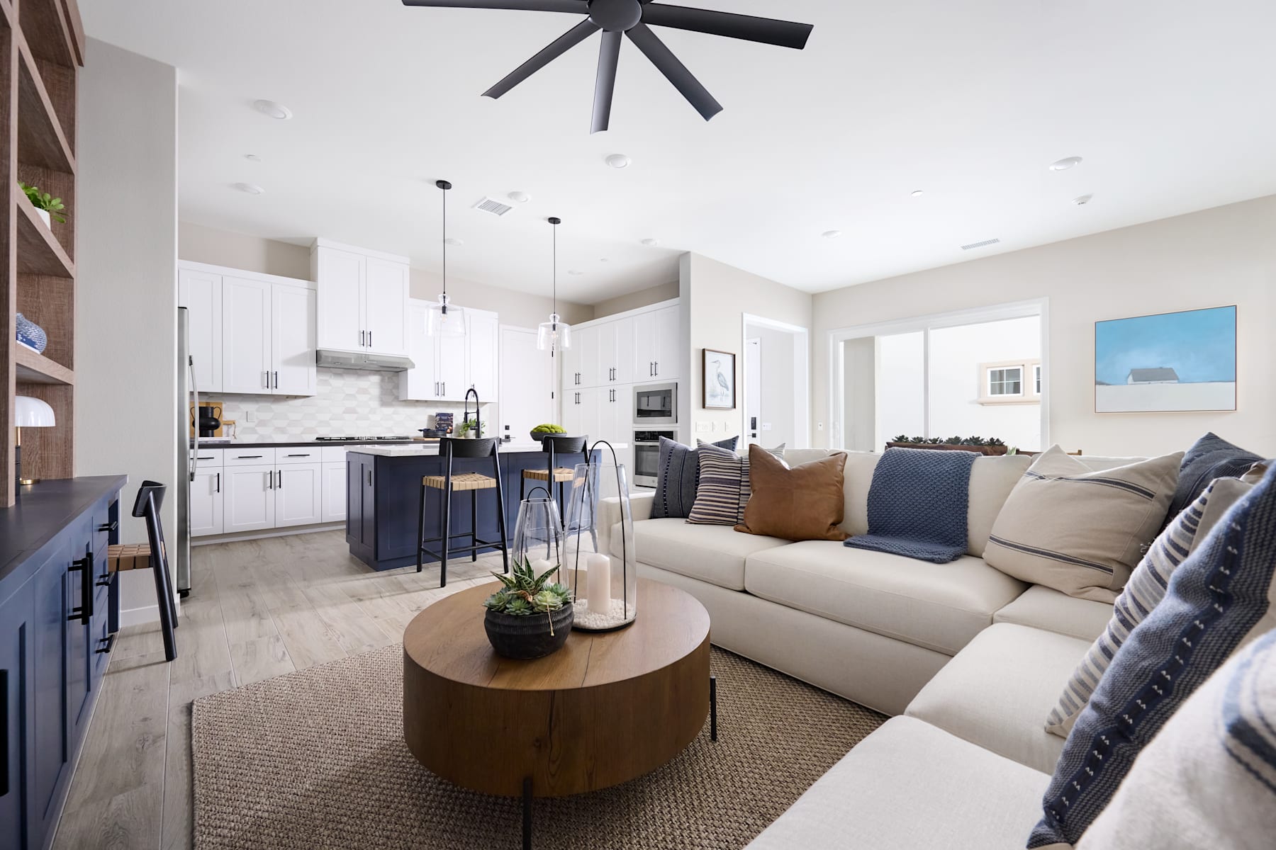 A modern and cozy open-concept living space with a white sectional sofa, a round wooden coffee table, and a large ceiling fan in the background.