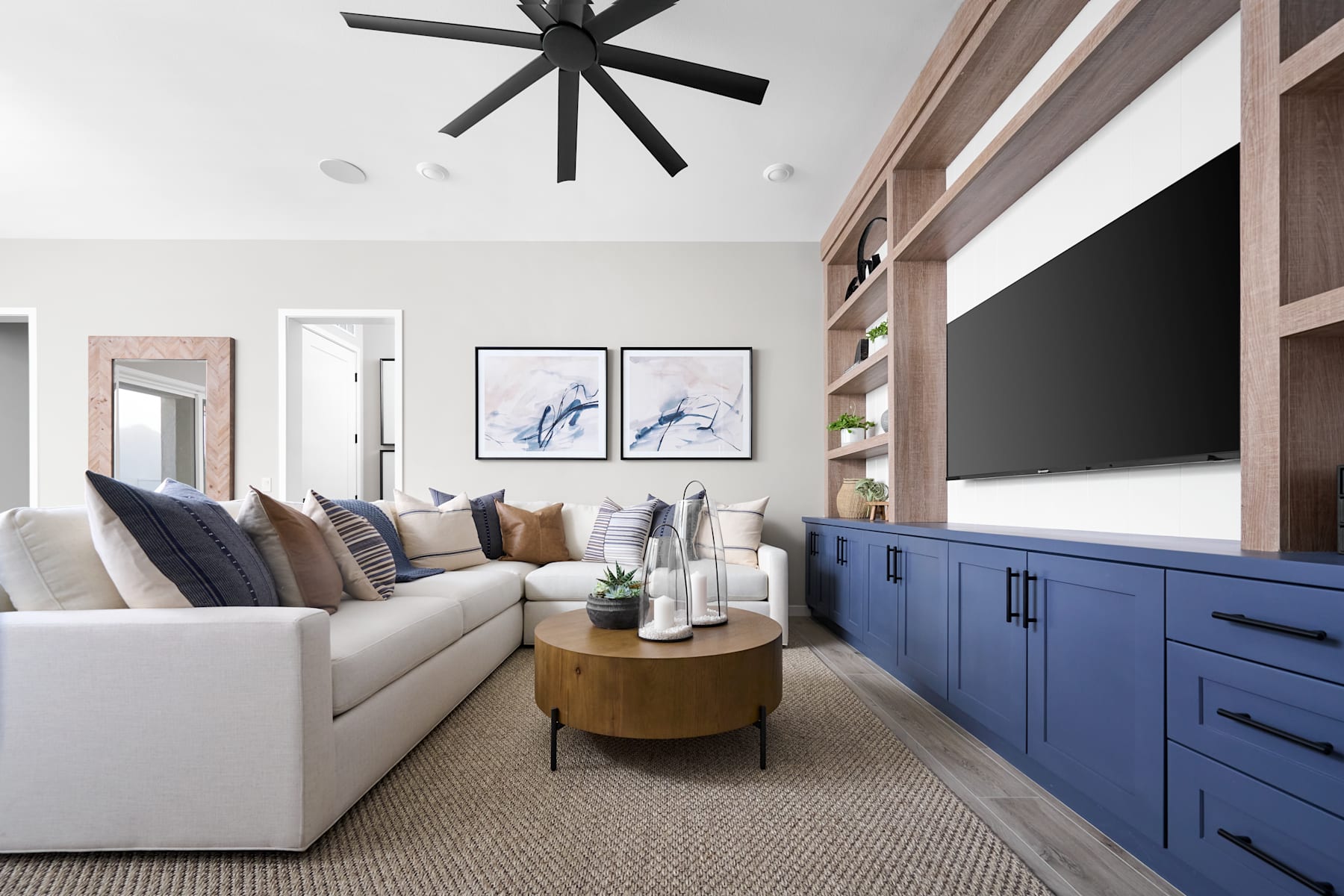 A modern and stylish living room with a white sectional sofa, a wooden coffee table, and a large black ceiling fan. The room features blue built-in cabinets and shelves, as well as framed artwork on the walls.