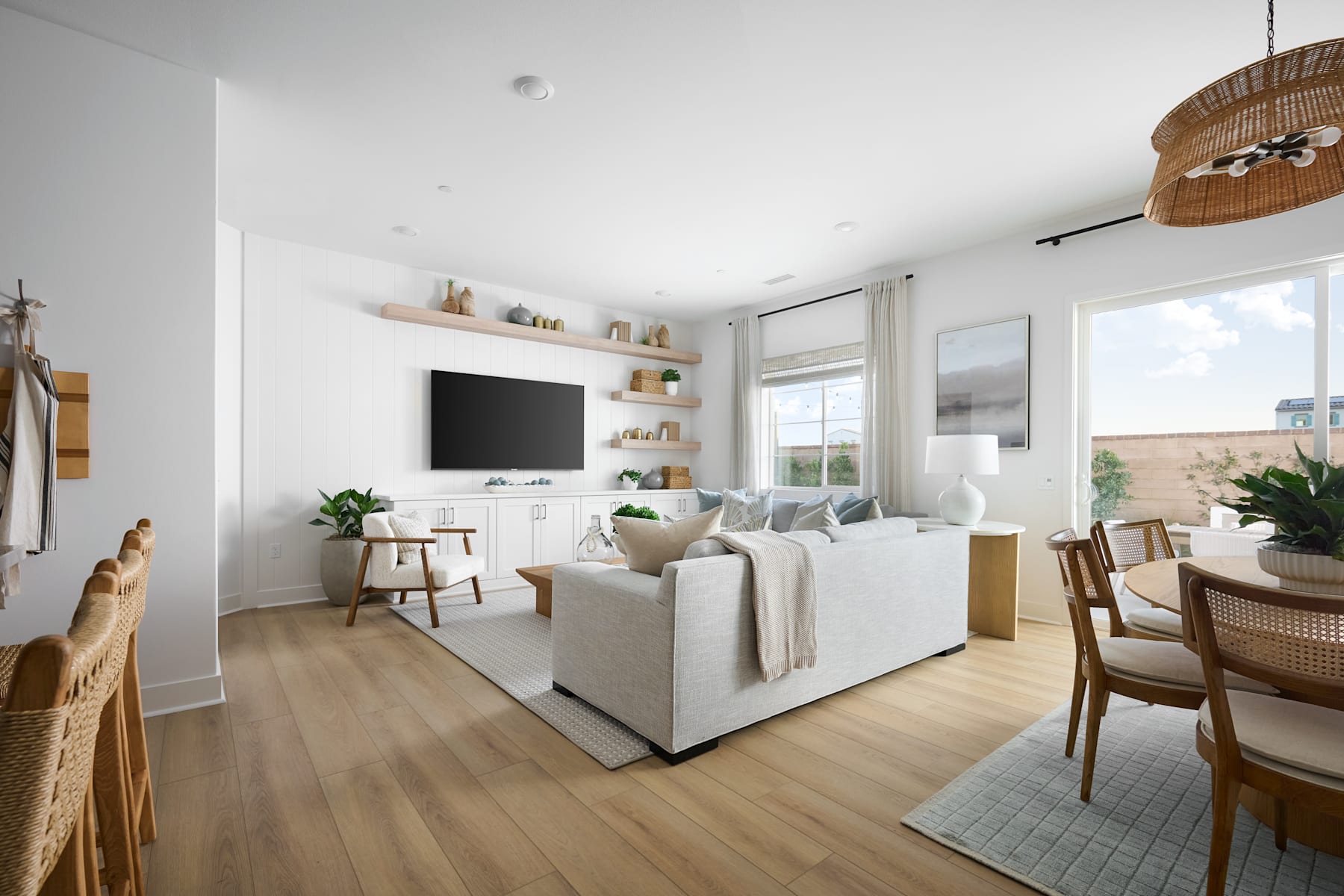 A bright and airy living room with a cozy seating area, a dining table, and shelves displaying decorative items, all set against a backdrop of large windows and natural light.