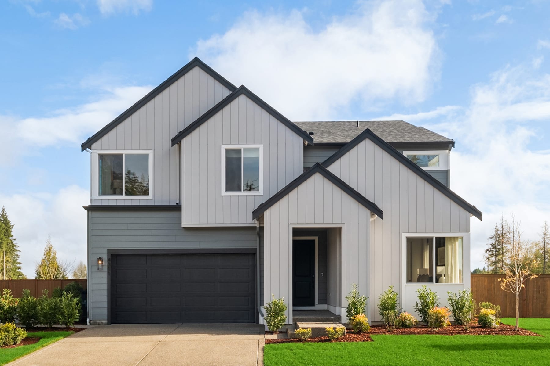 A modern two-story house with a gray exterior, a black garage door, and a well-manicured lawn in the foreground, set against a backdrop of blue sky with fluffy white clouds.