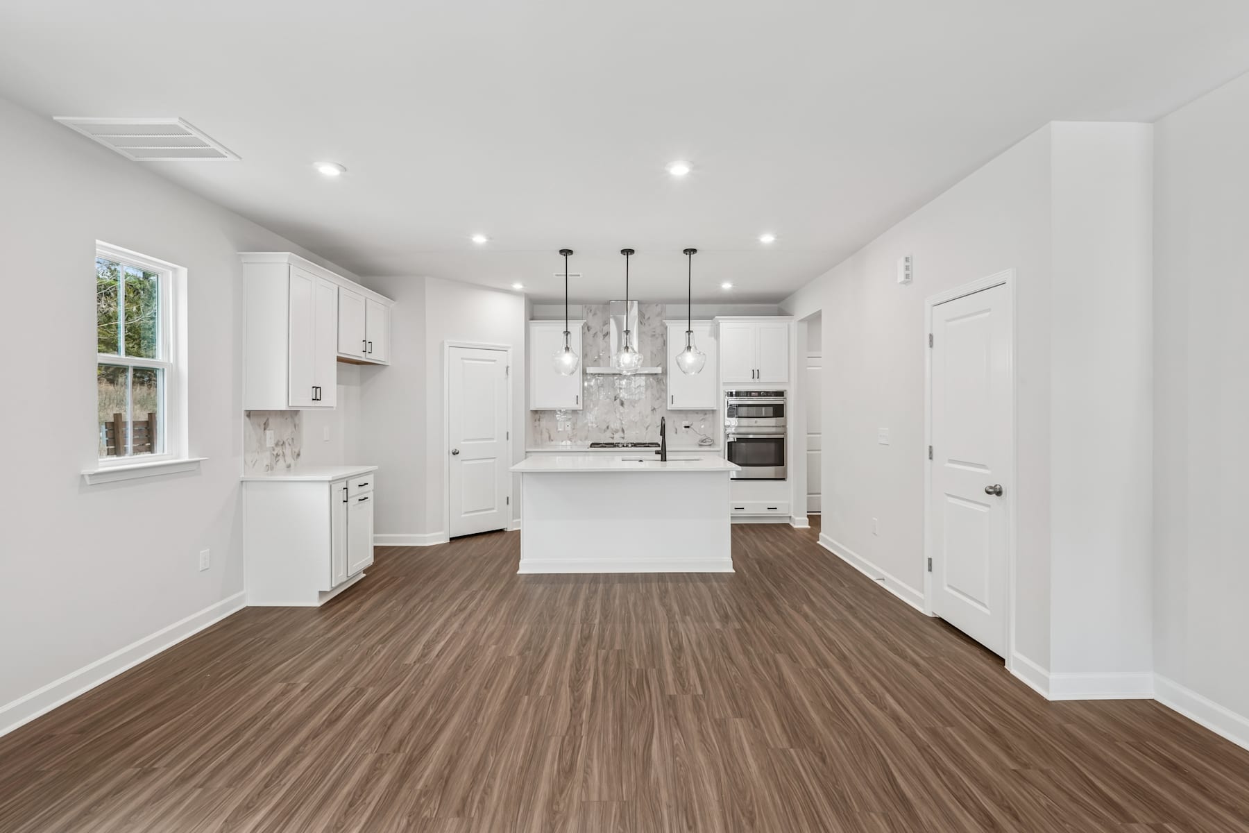 A bright, open-concept kitchen and living space with white cabinets, hardwood floors, and large windows providing natural light.