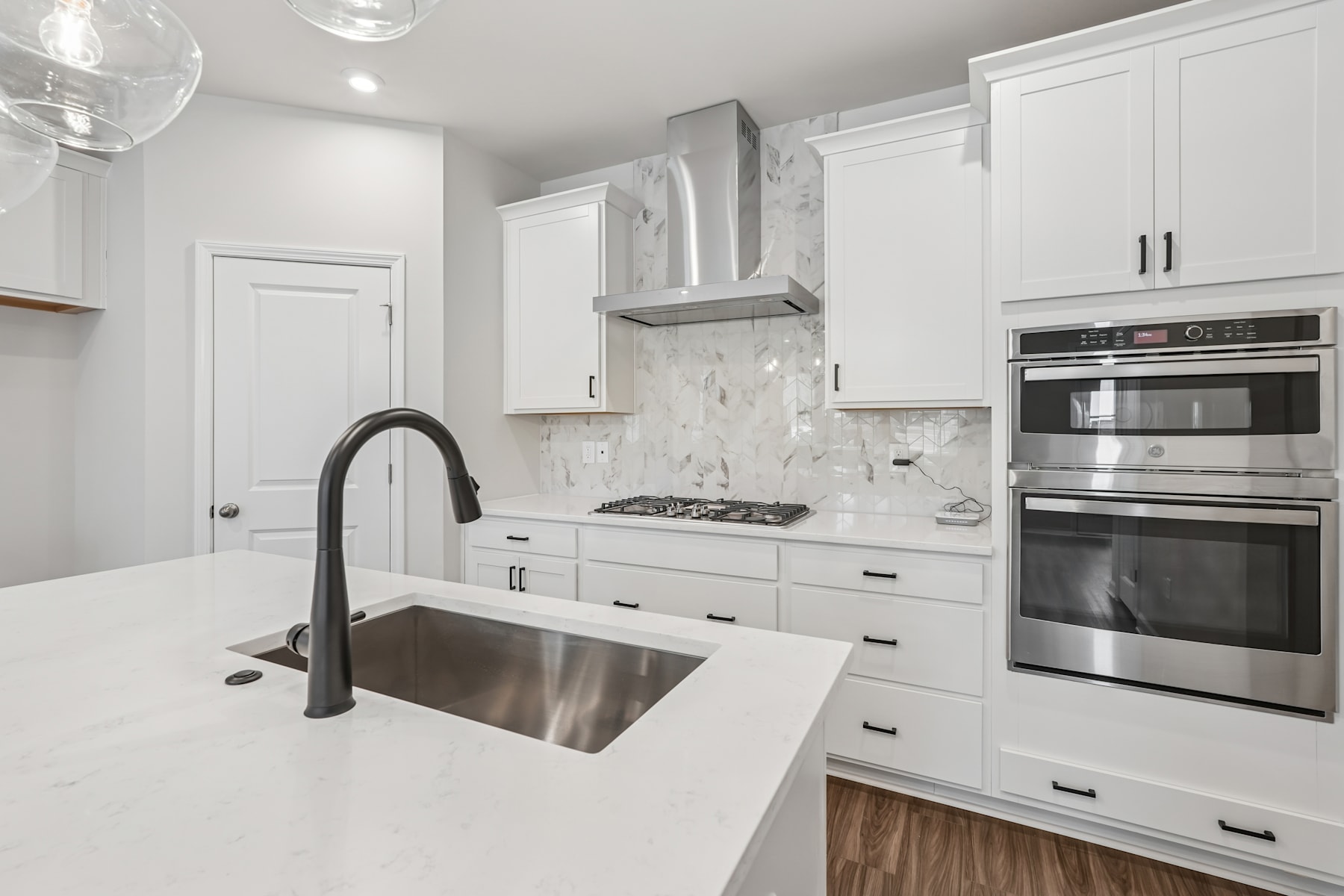 A modern, white kitchen with stainless steel appliances, a marble backsplash, and a large sink with a black faucet.
