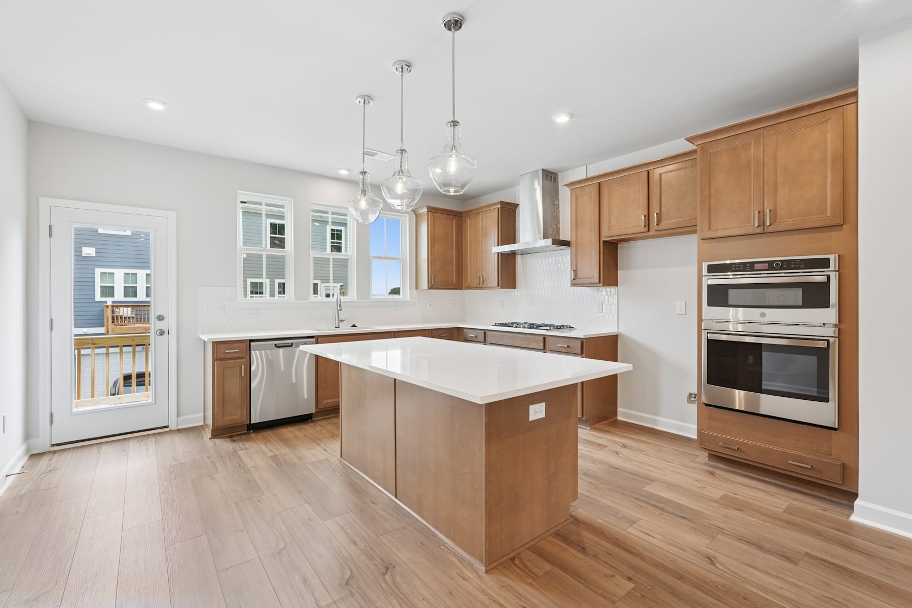 A modern, open-concept kitchen with light wood cabinetry, a central island, and stainless steel appliances, set against a backdrop of white walls and hardwood floors.