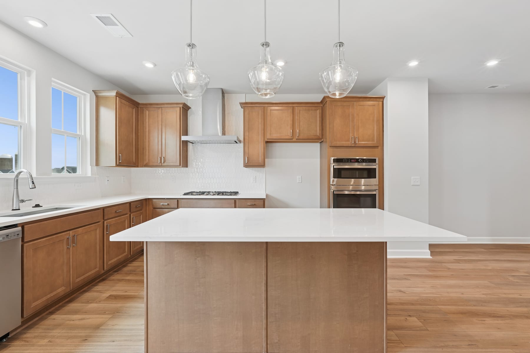 A modern, open-concept kitchen with light wood cabinetry, a large central island, and pendant lighting fixtures hanging above.