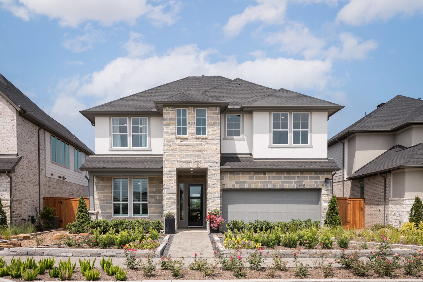 A two-story residential house with a stone exterior, a tiled roof, and a well-manicured front yard featuring various plants and flowers.