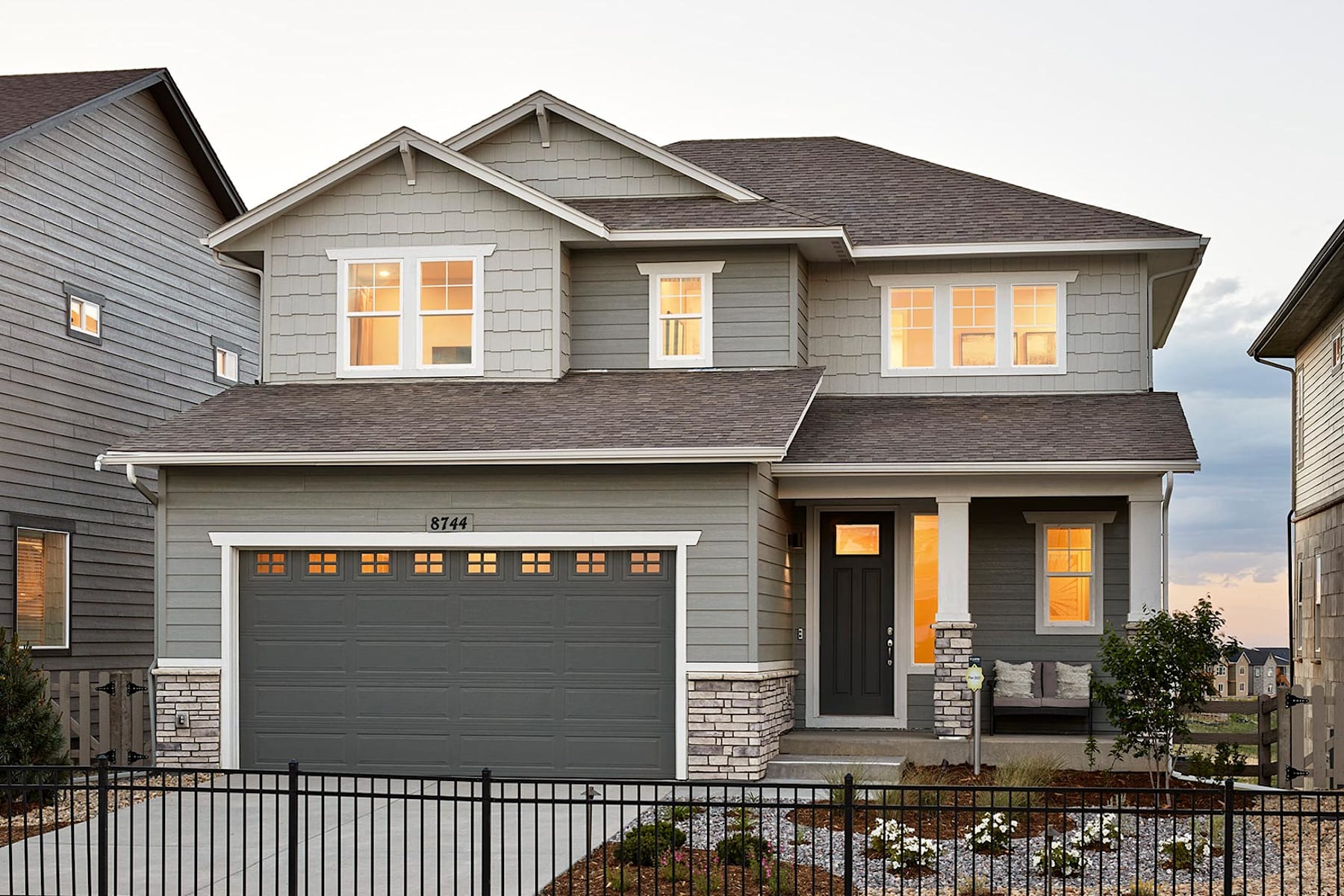 A two-story residential house with a gray exterior, a garage door, and illuminated windows, surrounded by a landscaped yard with flowers in the foreground.