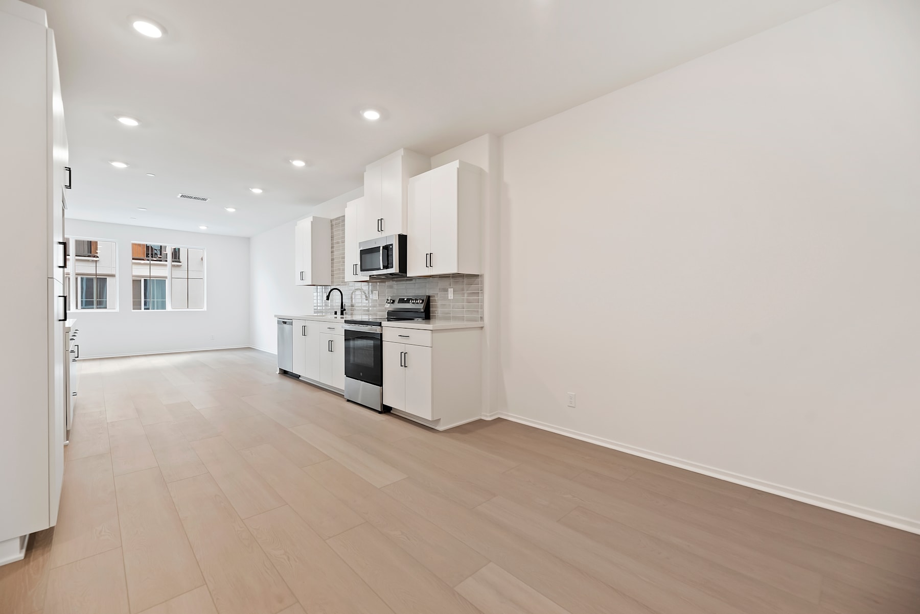 A modern, open-concept kitchen with white cabinets, stainless steel appliances, and a hardwood floor leading into a bright, airy living space.