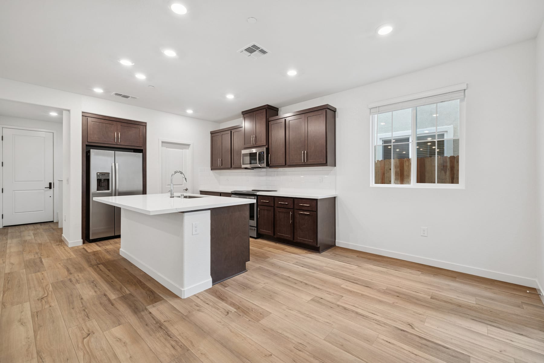 A modern, open-concept kitchen with dark wood cabinets, a white countertop, and hardwood floors, illuminated by recessed lighting.