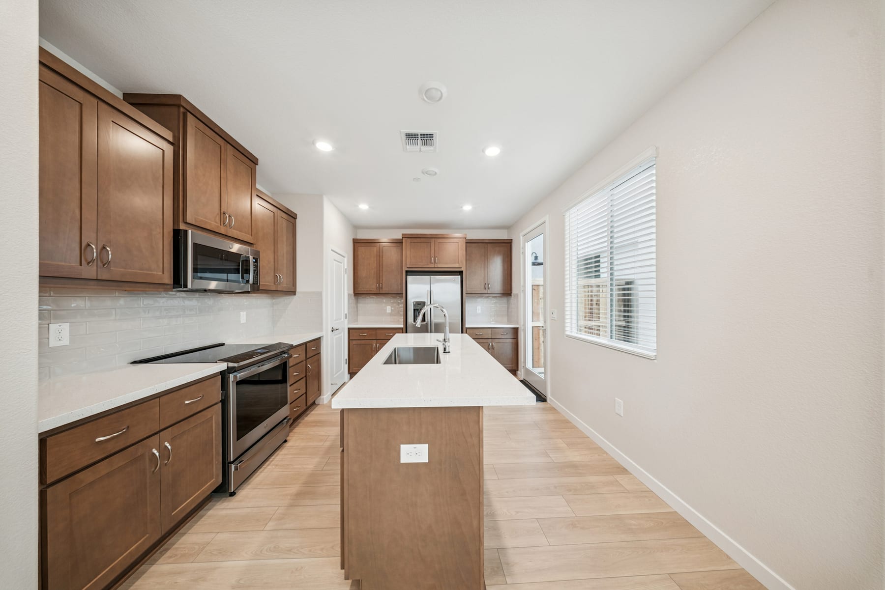 A modern, well-lit kitchen with wooden cabinets, stainless steel appliances, and a long central island with a white countertop.