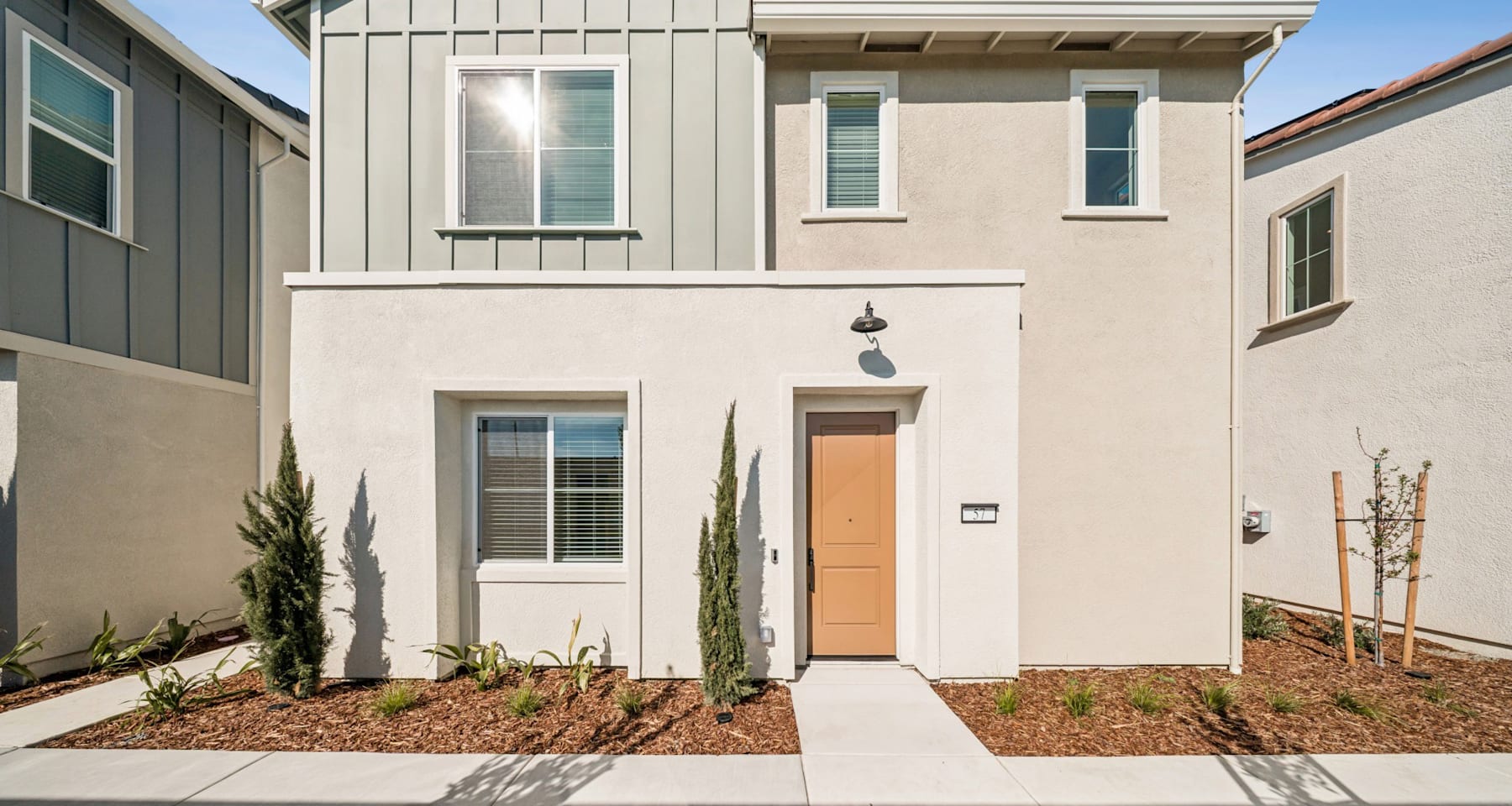 A modern, two-story residential building with a beige exterior, a bright orange front door, and a well-landscaped yard with various plants and shrubs in the foreground.
