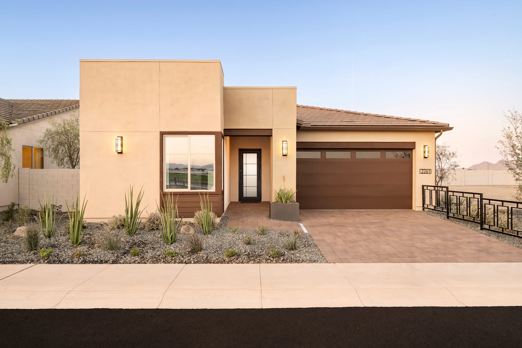A modern, single-story house with a beige exterior, a garage door, and a landscaped front yard with desert plants against a clear blue sky.