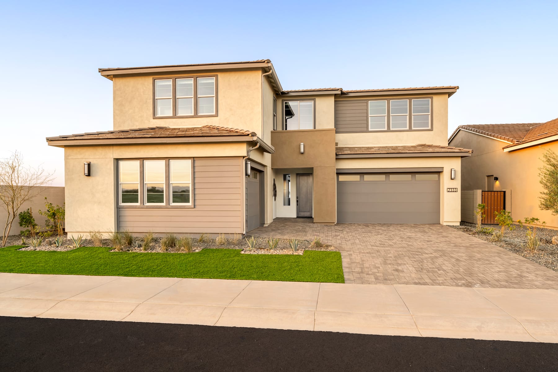 A two-story beige house with a garage, surrounded by a well-manicured lawn and a paved driveway, set against a clear blue sky.