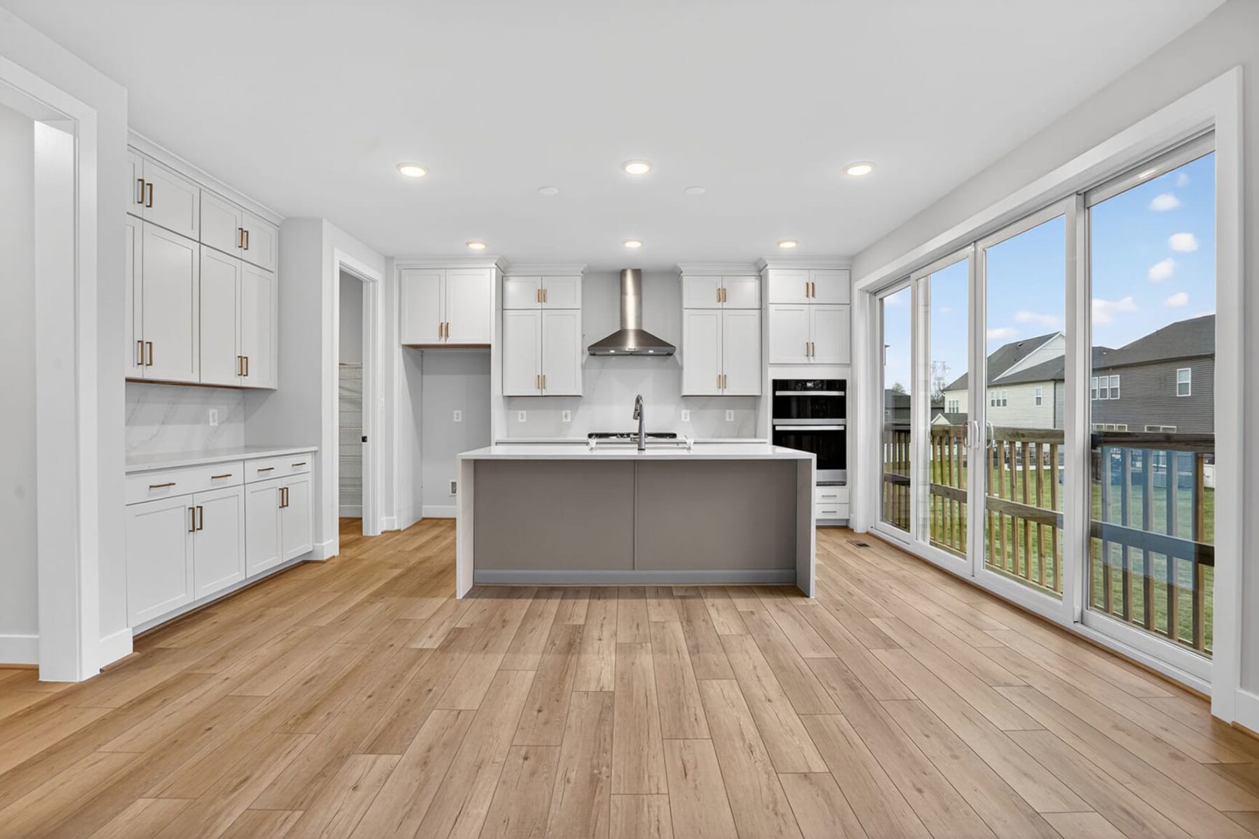 A modern, bright kitchen with white cabinets, a gray island, and large windows overlooking a backyard.