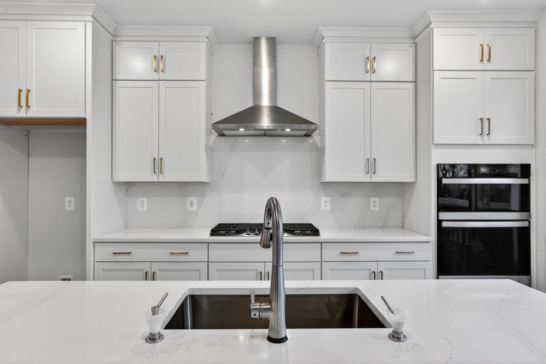 A modern, white kitchen with stainless steel appliances, including a range hood and a double oven, along with a large sink and countertop in the foreground.
