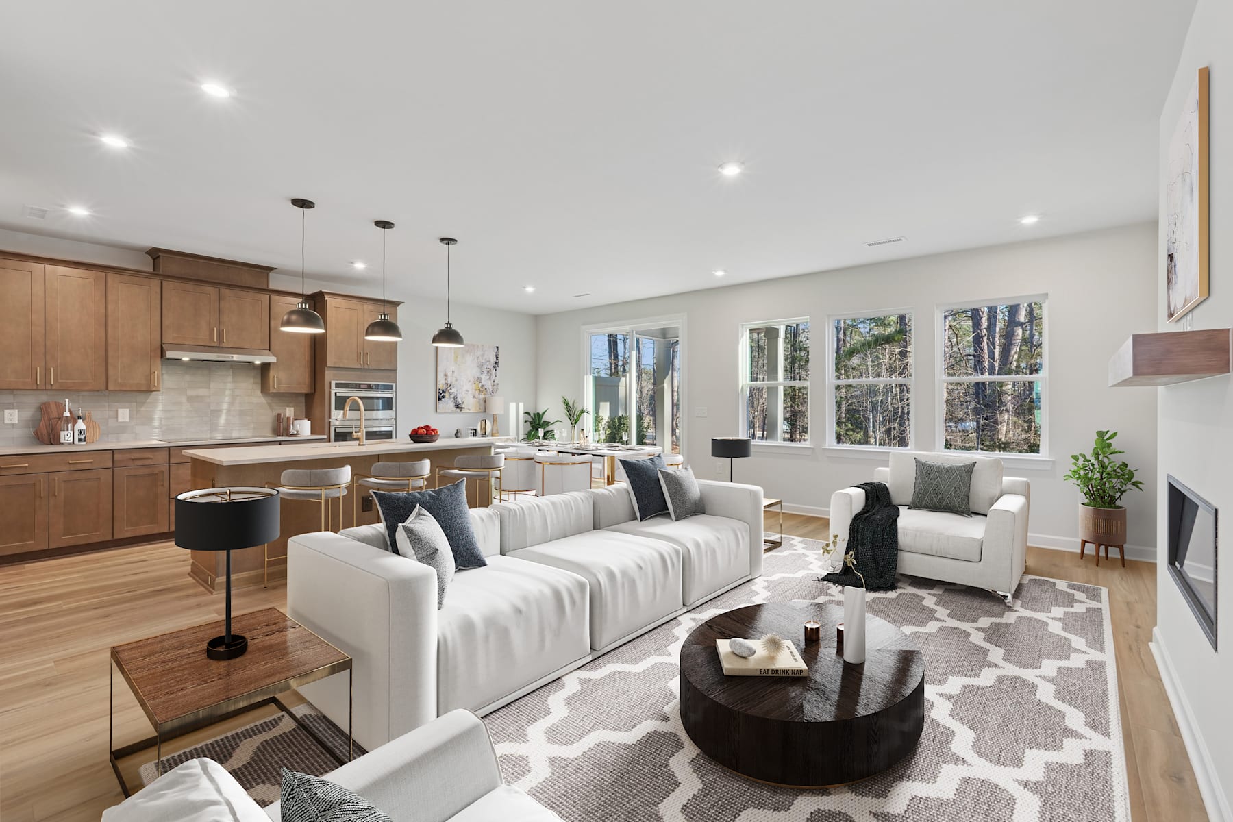 A modern and spacious open-concept living room and kitchen with wooden cabinets, a large couch, and a patterned rug in the foreground, surrounded by large windows overlooking a wooded outdoor area.