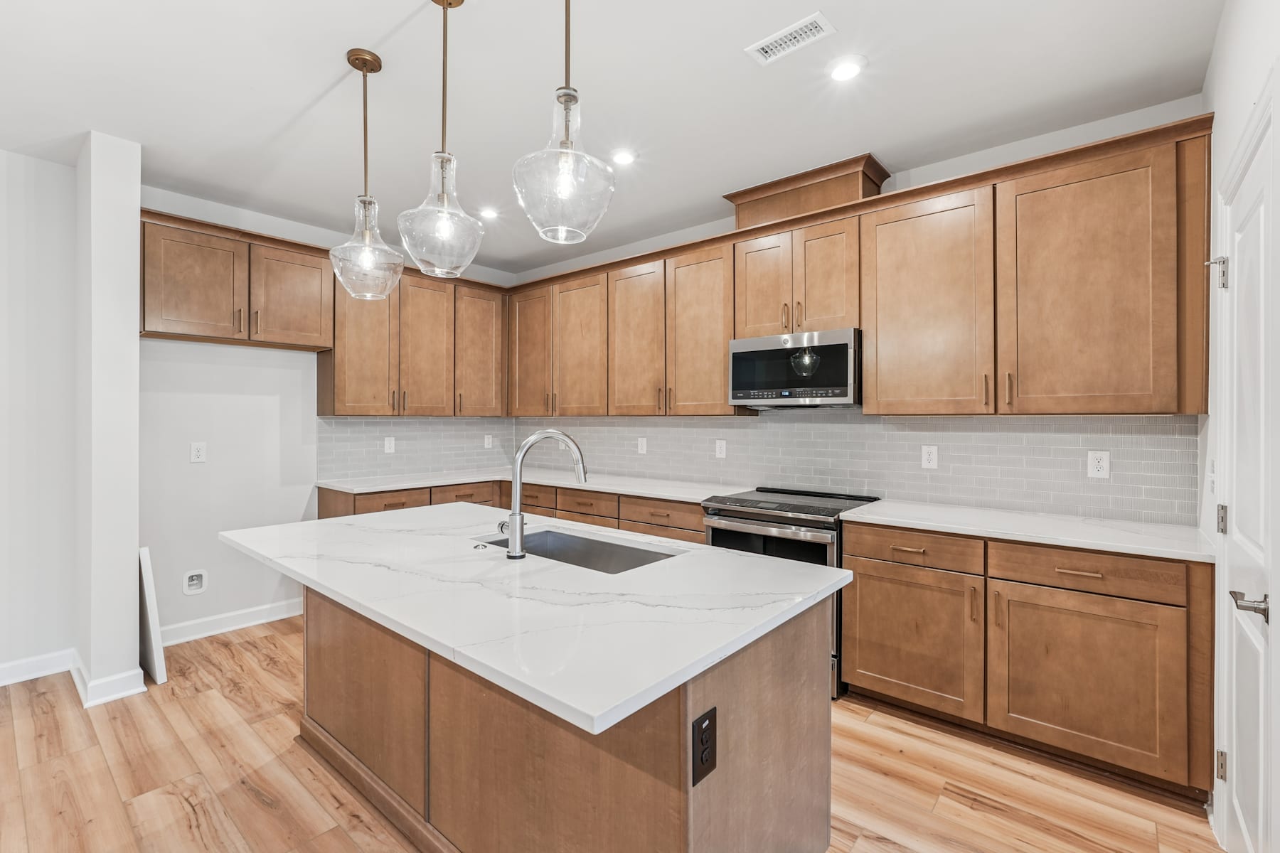 A modern kitchen with wooden cabinets, a white countertop, and pendant lighting fixtures, set against a bright and airy background.
