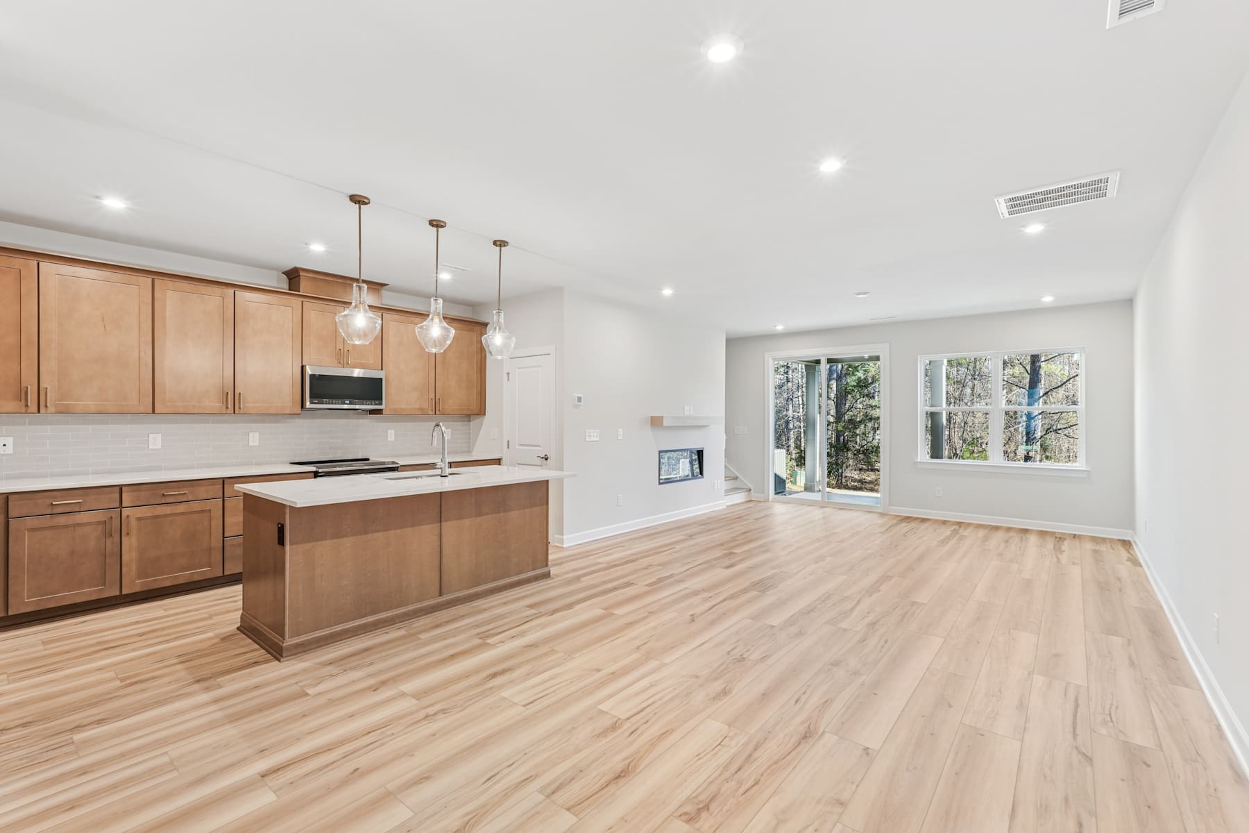 A modern, open-concept kitchen and living room with wooden cabinets, hardwood floors, and large windows providing natural light.
