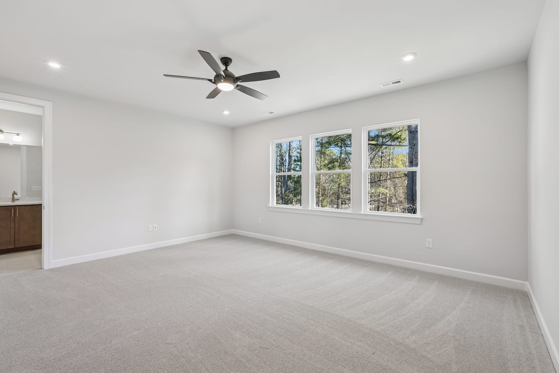 A spacious, well-lit room with a ceiling fan, large windows overlooking a wooded area, and a clean, carpeted floor.