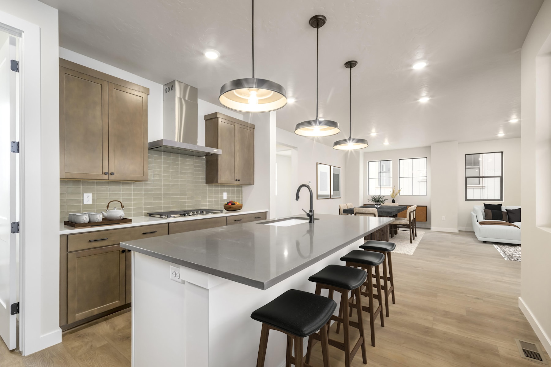 A modern and spacious kitchen with light-colored cabinets, a large island with bar stools, and pendant lights hanging from the ceiling, leading into an open living area with a couch and windows.
