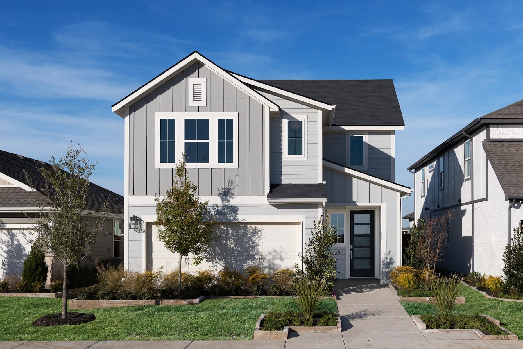 A two-story residential house with a gabled roof, gray siding, and black-framed windows stands in a well-manicured yard with trees and shrubs in the foreground against a clear blue sky.