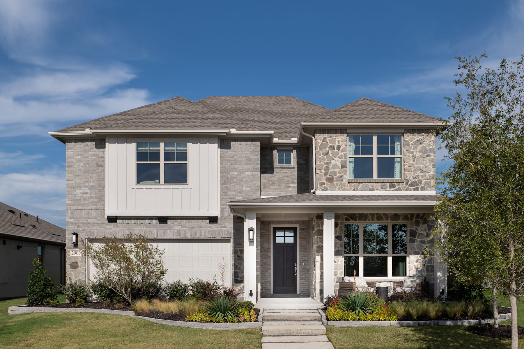 A two-story residential house with a stone and stucco exterior, surrounded by a well-manicured lawn and landscaping, set against a clear blue sky with wispy clouds.