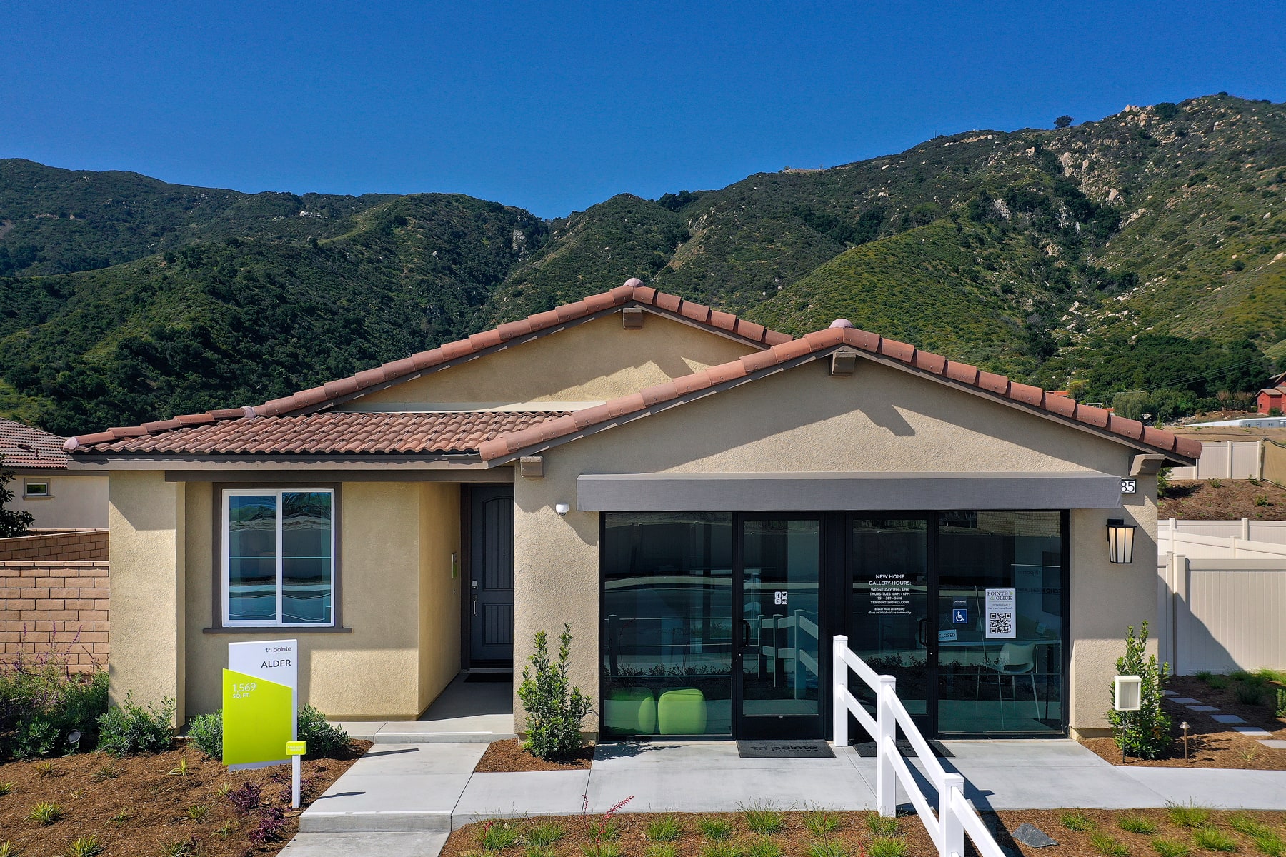 A single-story house with a tiled roof sits in the foreground, surrounded by lush greenery and rugged mountains in the background.