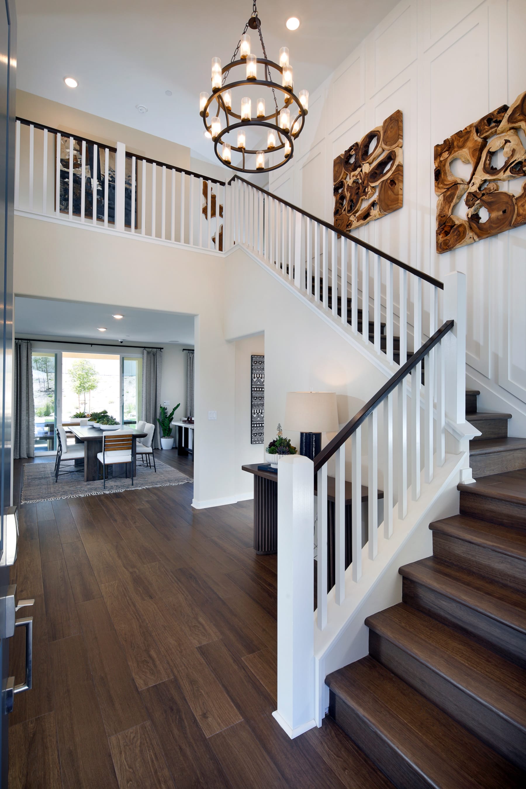 A spacious and well-lit foyer with a grand staircase, wooden floors, and a decorative chandelier, leading to a living area with a desk and artwork on the walls.