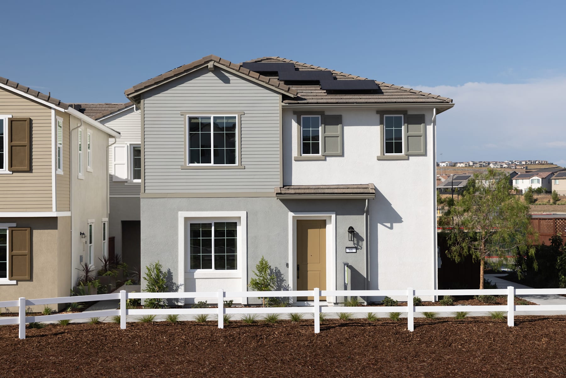 A two-story residential house with a white exterior, a slanted roof, and a white picket fence in the foreground, set against a backdrop of other houses and a clear blue sky.