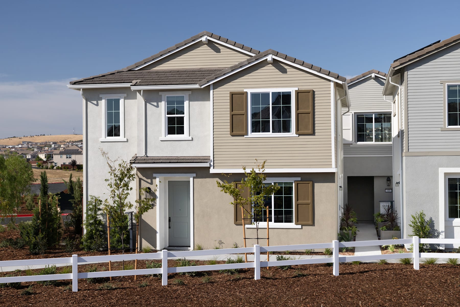 A two-story residential building with a white picket fence in the foreground, surrounded by landscaped greenery and a clear blue sky in the background.