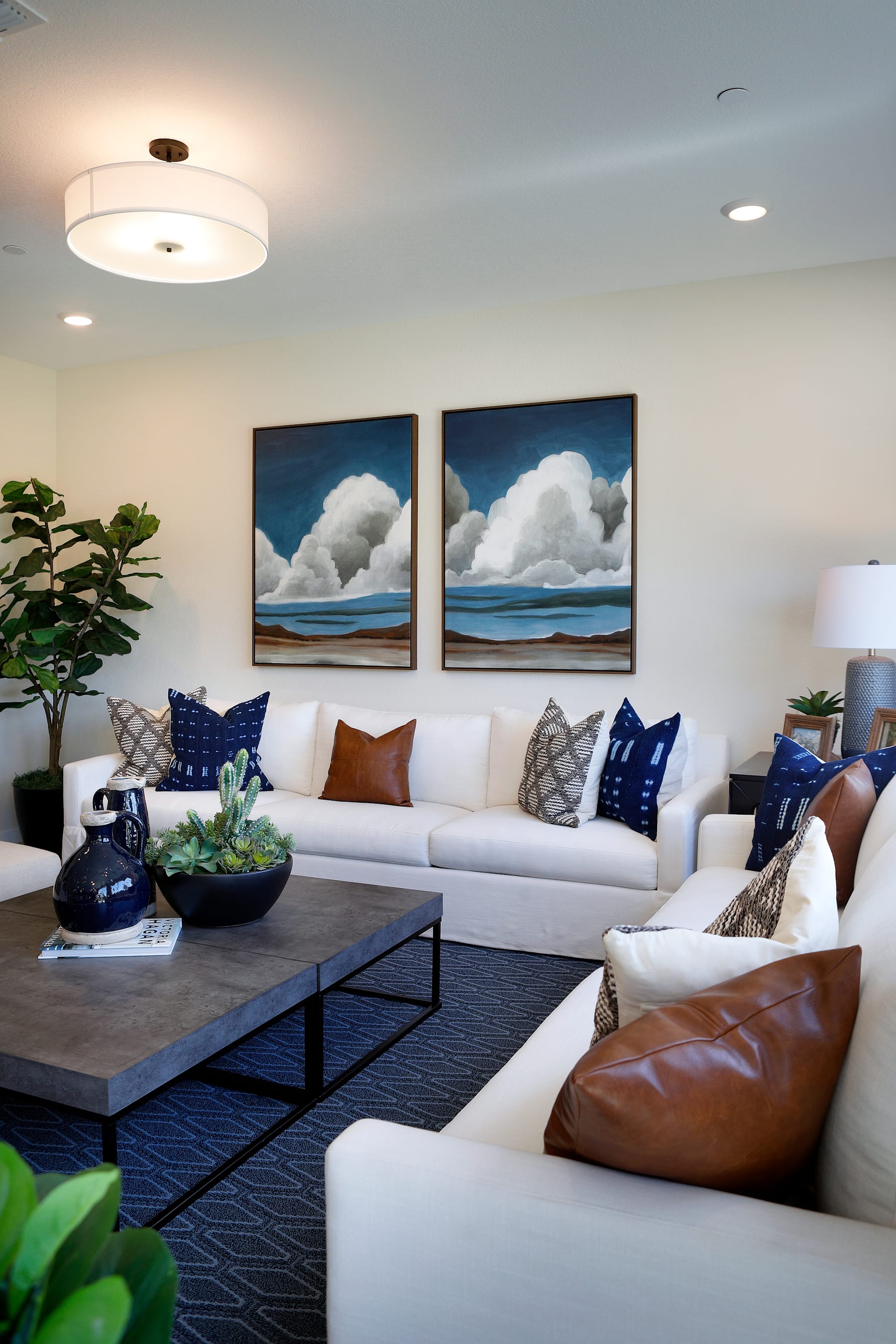 A modern and cozy living room with a large white sofa, a leather armchair, and two framed paintings of cloudy skies and a body of water on the walls.