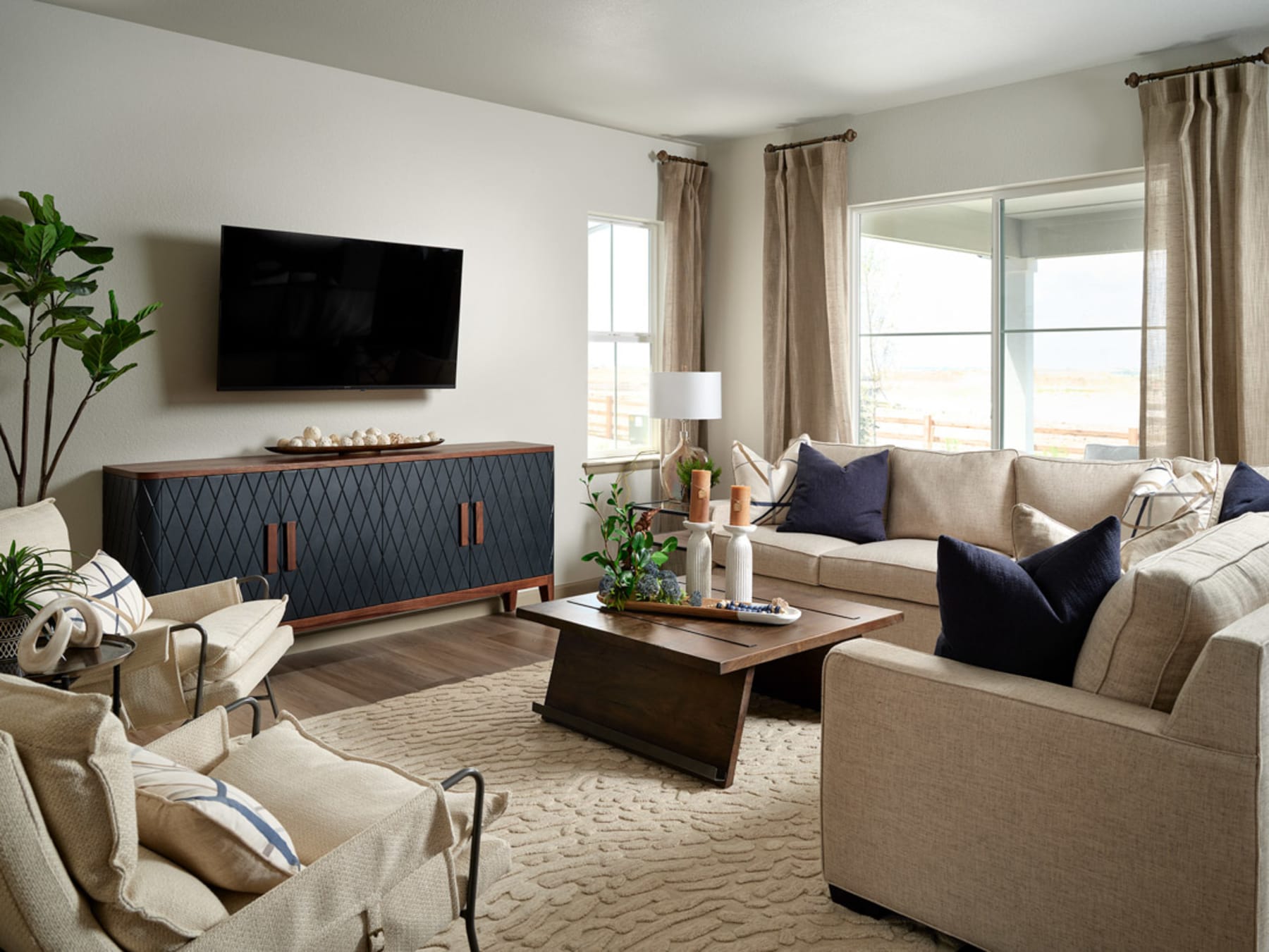 A cozy and well-furnished living room with a large TV, a wooden cabinet, and a comfortable seating arrangement featuring beige and navy blue sofas, along with various decorative elements such as plants and lamps.