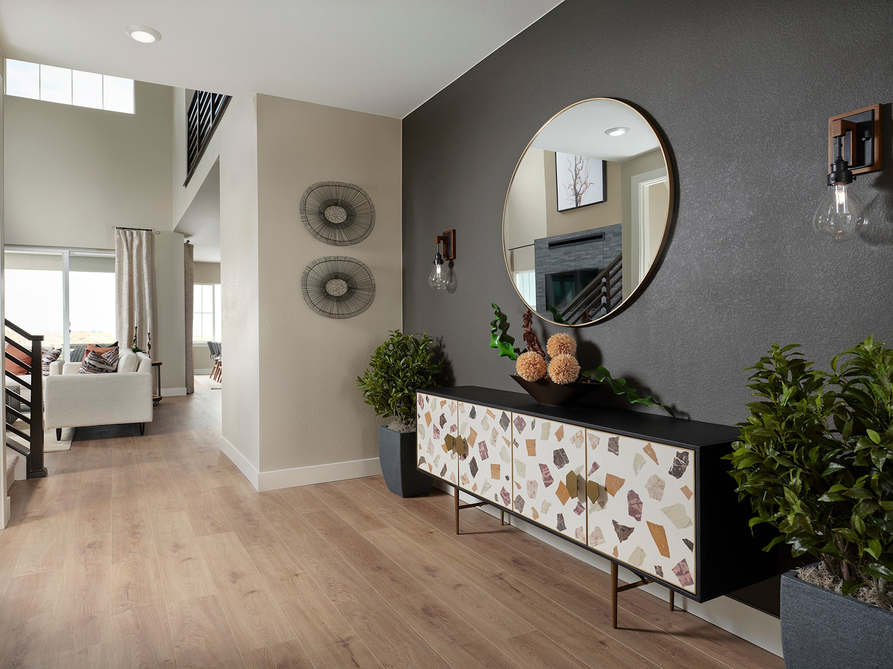 A modern and stylish entryway with a dark-colored accent wall, a round mirror, and a decorative console table with a mosaic pattern, surrounded by potted plants and other decor elements.
