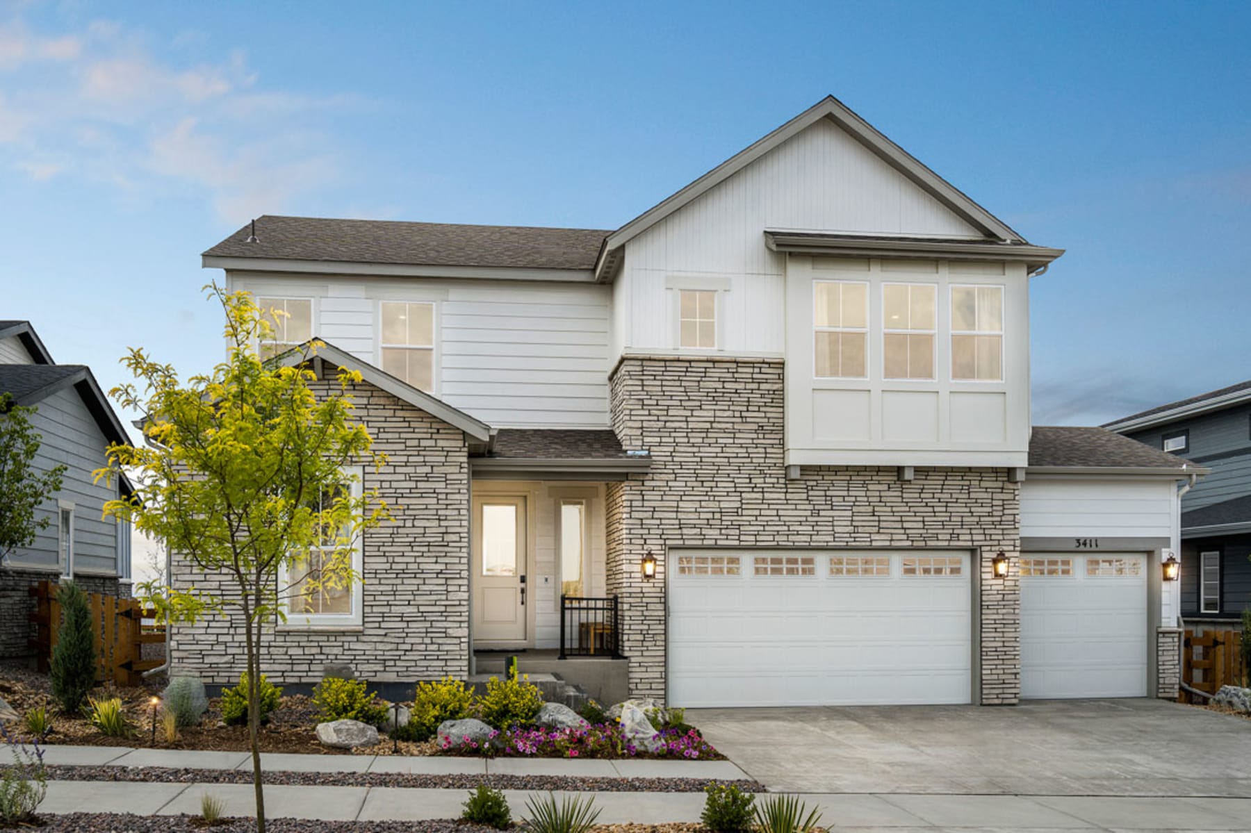 A two-story residential house with a white exterior, stone accents, and a garage door in the foreground, set against a backdrop of a clear blue sky and lush greenery.