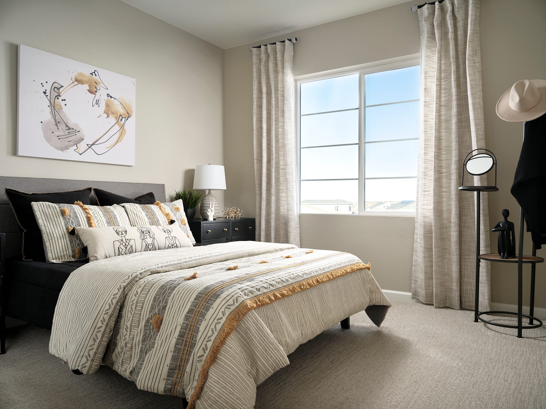 A cozy and well-decorated bedroom with a large window, a black bed frame, and a patterned bedspread, along with various decorative elements such as lamps and artwork on the walls.