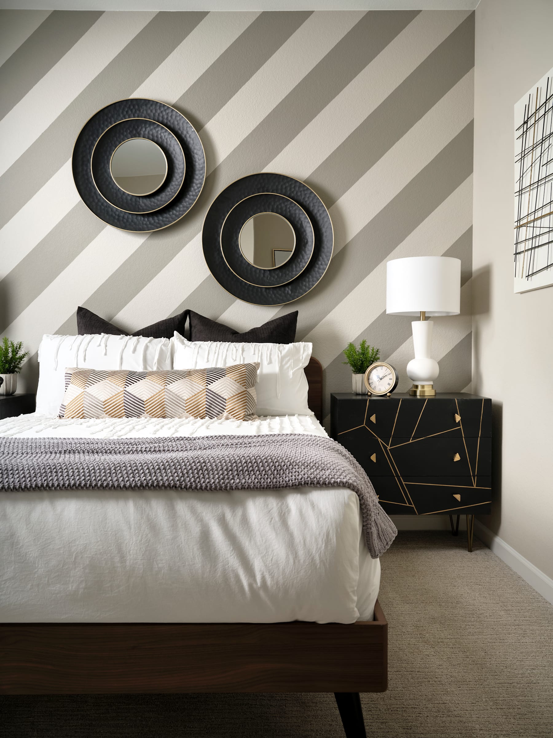 The image depicts a modern and stylish bedroom interior, featuring a large bed with a gray and white striped wall behind it, accented by two circular black and white wall decor pieces. A nightstand with a lamp and other decorative elements can be seen in the foreground.