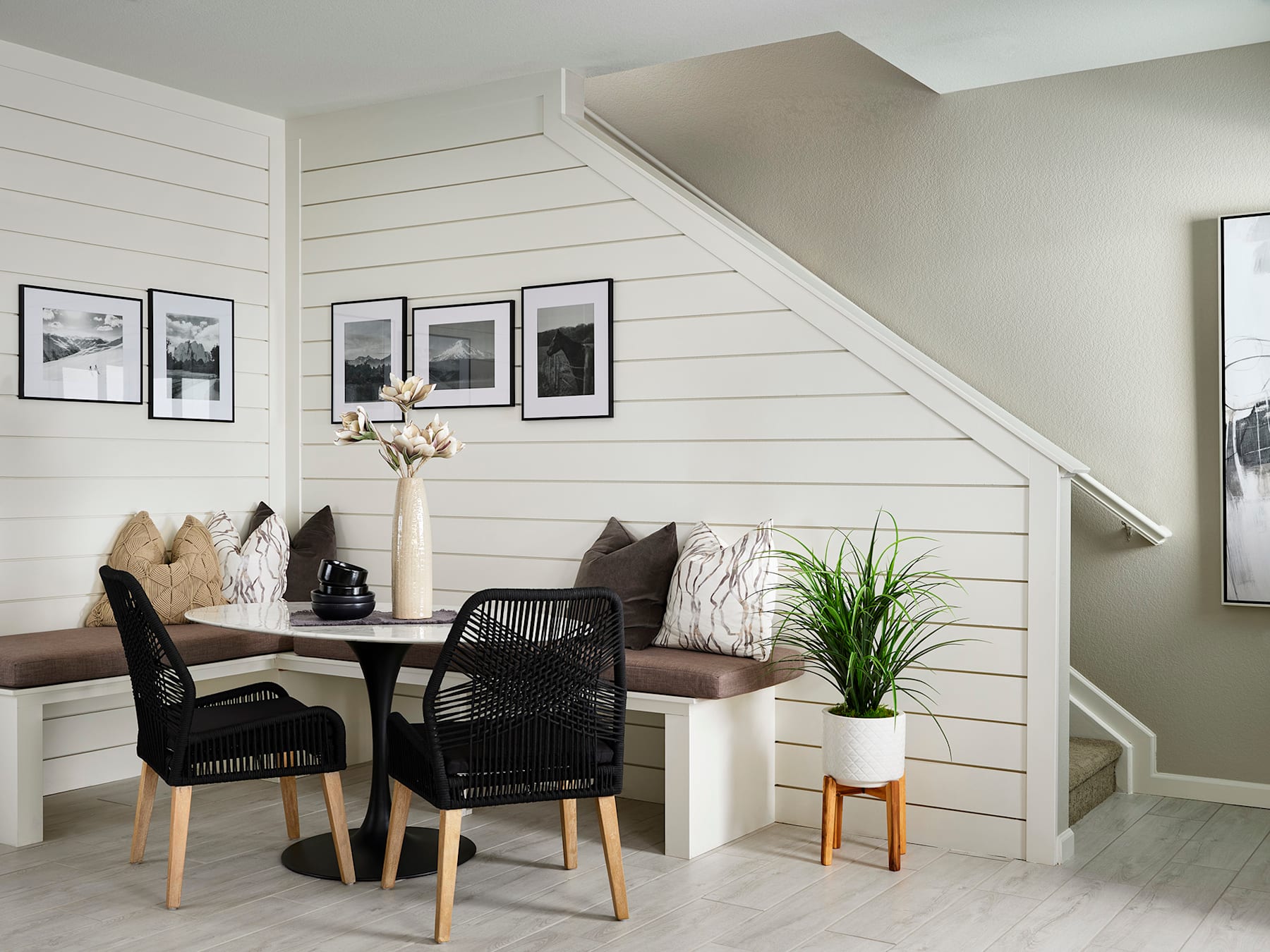 A cozy dining area with a wooden table, black wicker chairs, and framed artwork on the shiplap-style walls, complemented by a potted plant and a decorative vase.