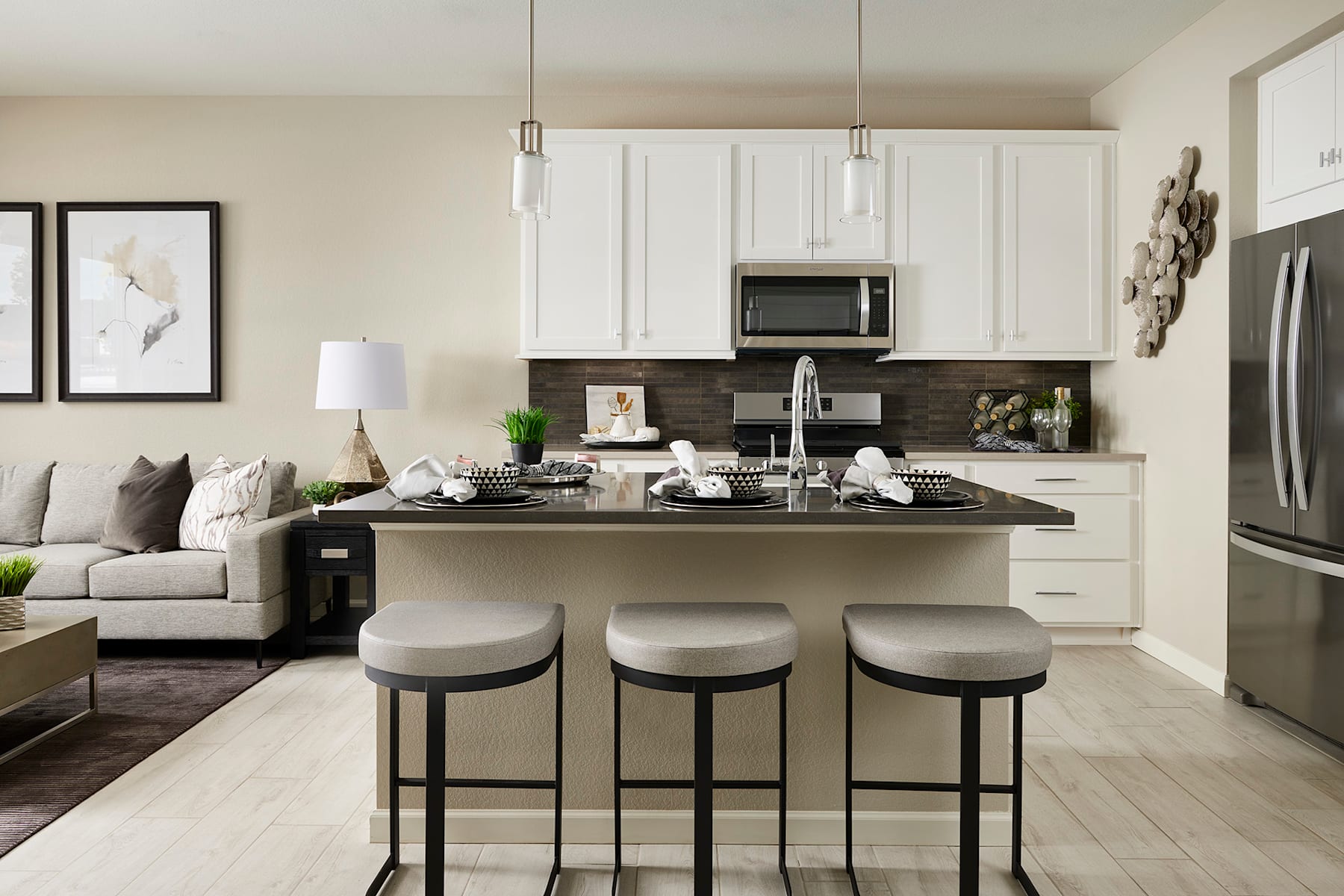 A modern and stylish kitchen with white cabinets, a dark countertop, and pendant lighting, situated in an open-concept living space with a gray sofa and framed artwork on the walls.