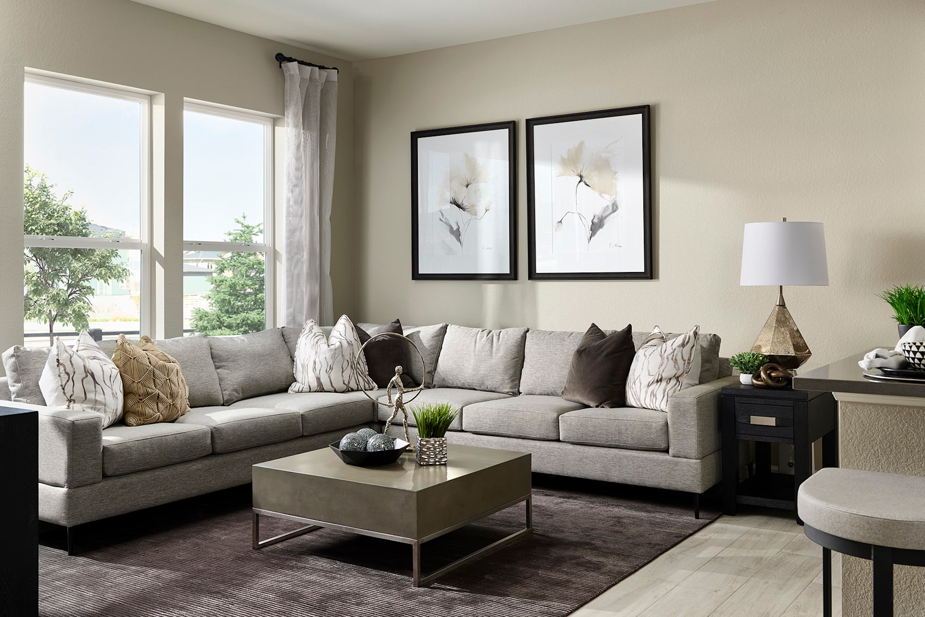 A cozy and inviting living room with a large gray sectional sofa, framed artwork on the walls, and a wooden coffee table in the foreground, surrounded by natural light from the windows in the background.