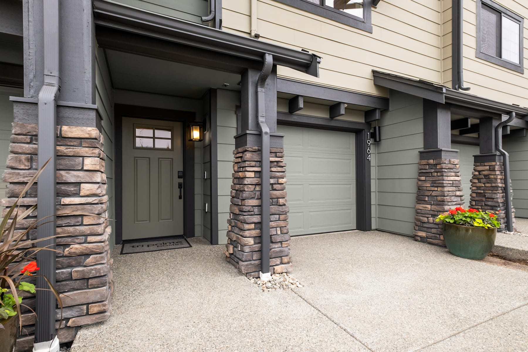 The image shows a modern, multi-unit residential building with a stone and siding exterior, a covered entryway, and a potted plant on the porch.