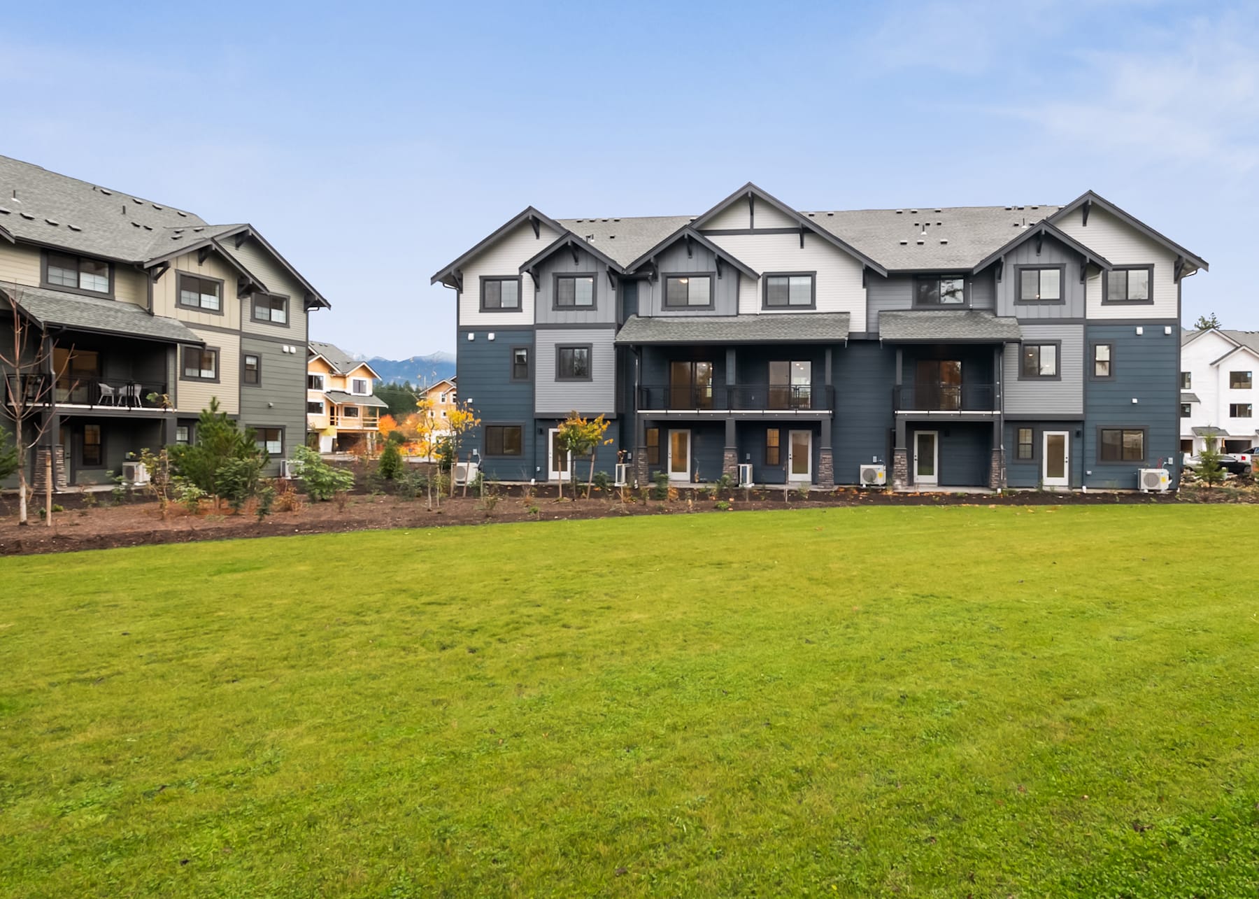 A modern multi-unit residential building with a grassy lawn in the foreground, set against a backdrop of mountains.
