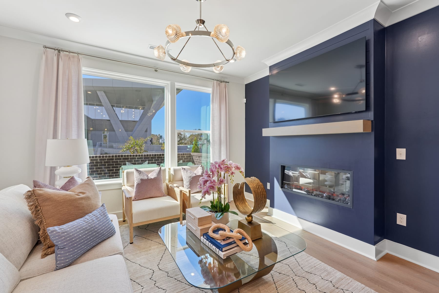 A cozy and well-decorated living room with a fireplace, comfortable seating, and a chandelier overhead, surrounded by large windows providing a scenic outdoor view.