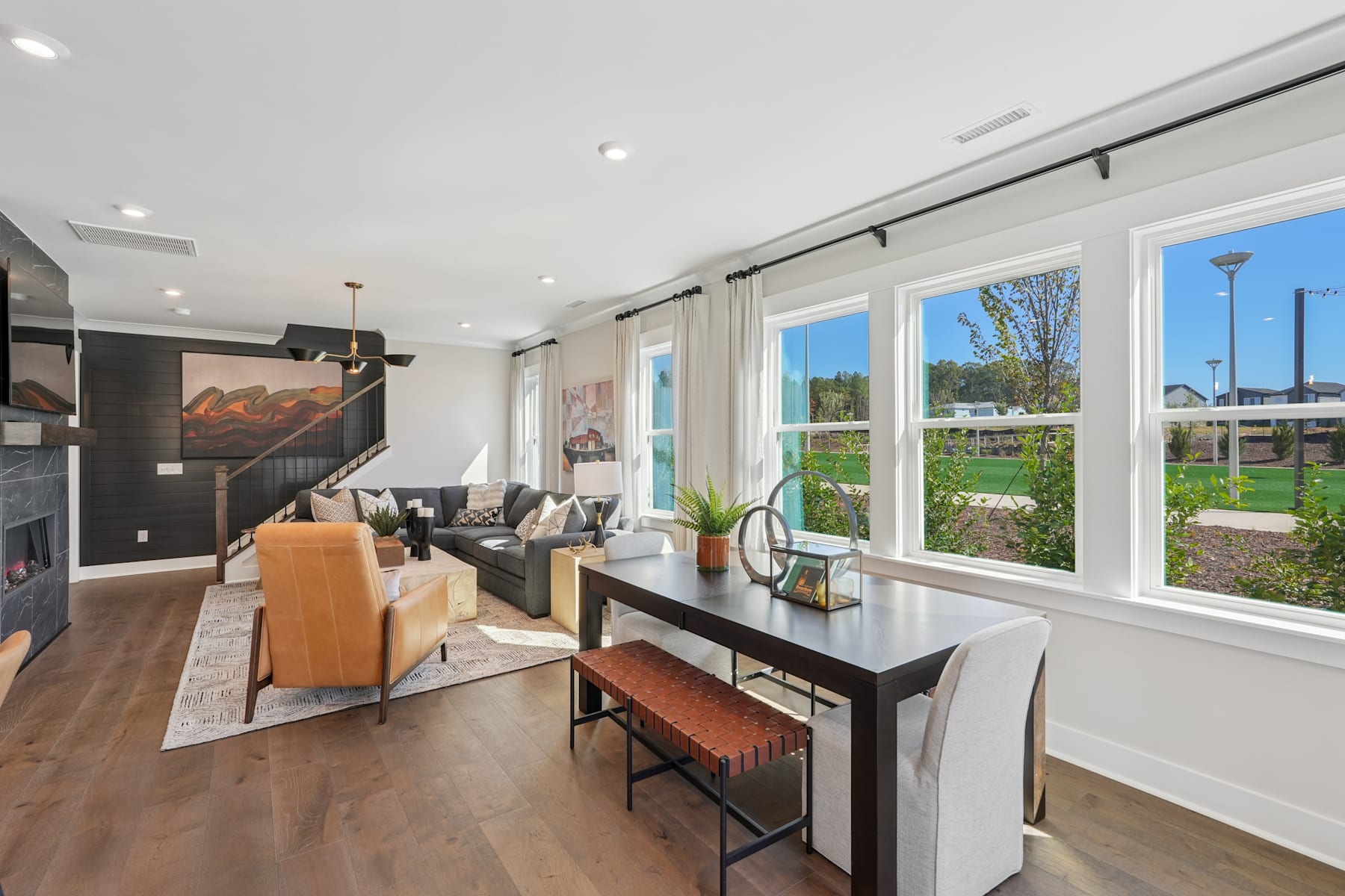 A modern and spacious living room with large windows overlooking a lush outdoor landscape, featuring a sleek black table, comfortable seating, and a warm, inviting atmosphere.