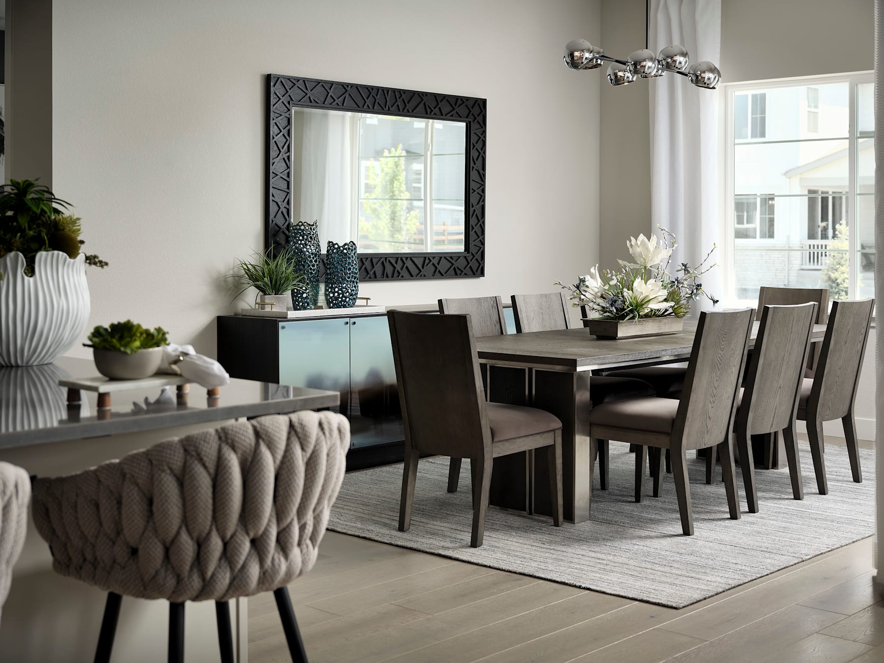 A modern and stylish dining room with a large wooden table, plush upholstered chairs, and a decorative mirror on the wall, complemented by potted plants and other decor elements.