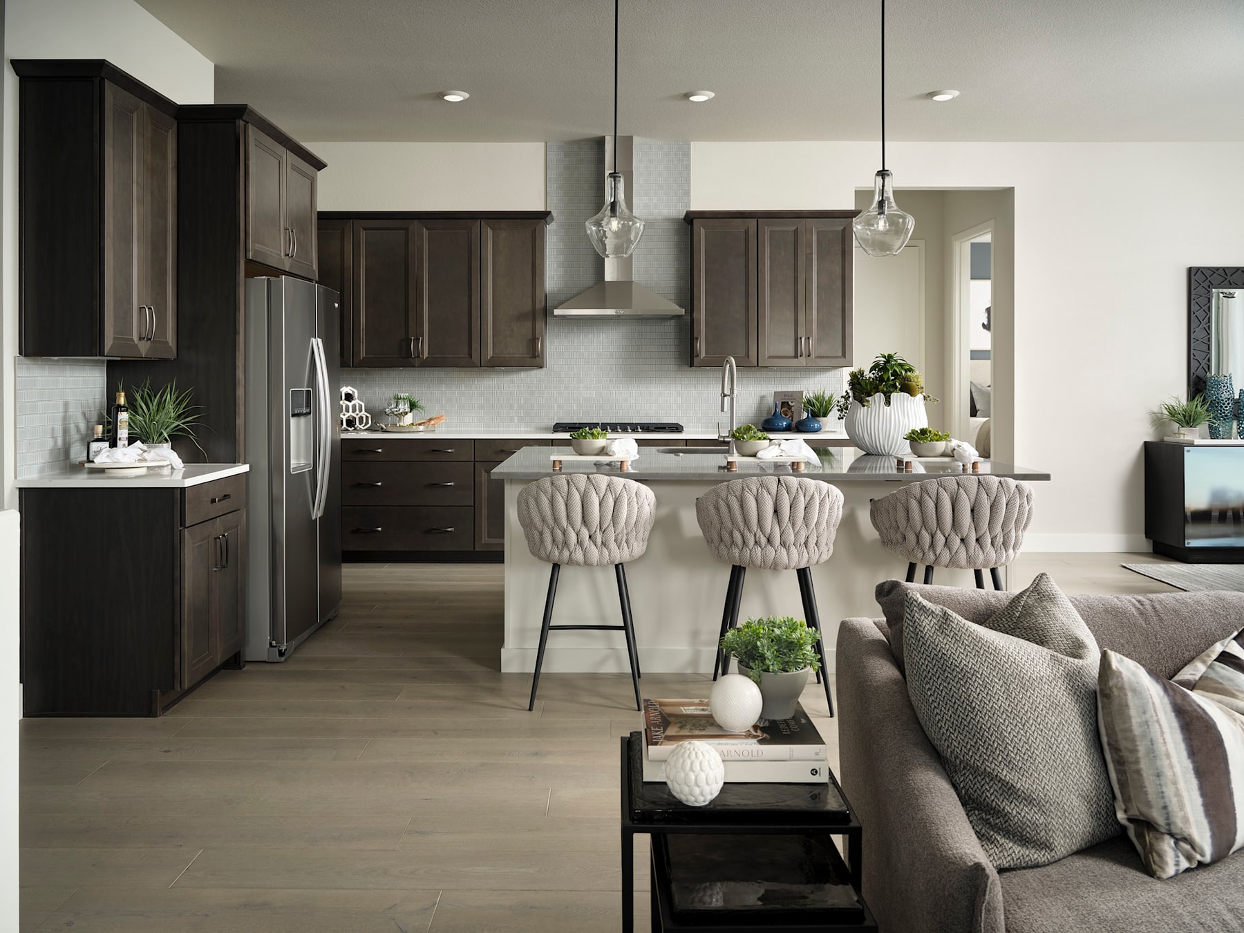 A modern and stylish kitchen with dark wood cabinets, a central island with plush upholstered stools, and a cozy living area with a gray sofa and decorative accents.