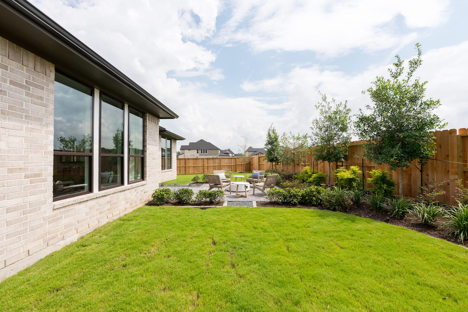A well-manicured backyard with a lush green lawn, surrounded by a wooden fence and featuring a patio area with outdoor furniture, set against a backdrop of a modern brick building with large windows.