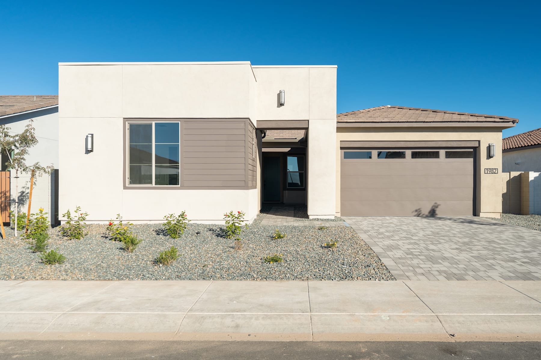 A modern, single-story house with a beige exterior, a garage, and a paved driveway in the foreground, set against a clear blue sky in the background.