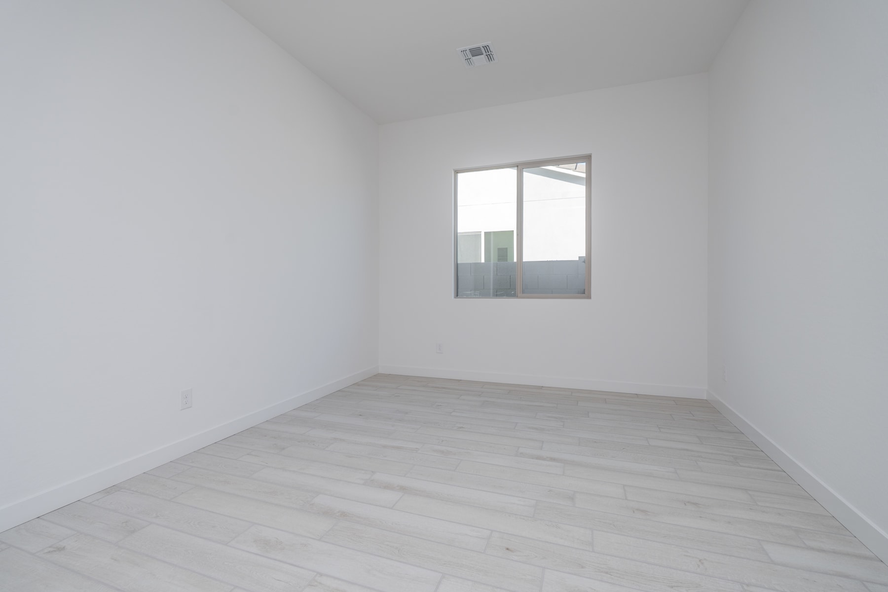 A minimalist, empty room with white walls and a wooden floor, featuring a single window providing natural light.