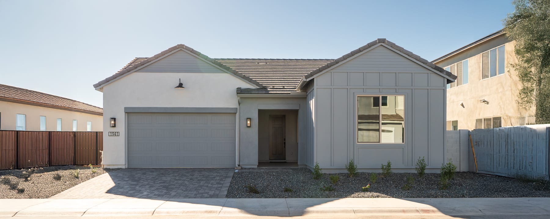 A modern, two-story house with a gray exterior, a garage, and a paved driveway in the foreground, set against a clear blue sky in the background.