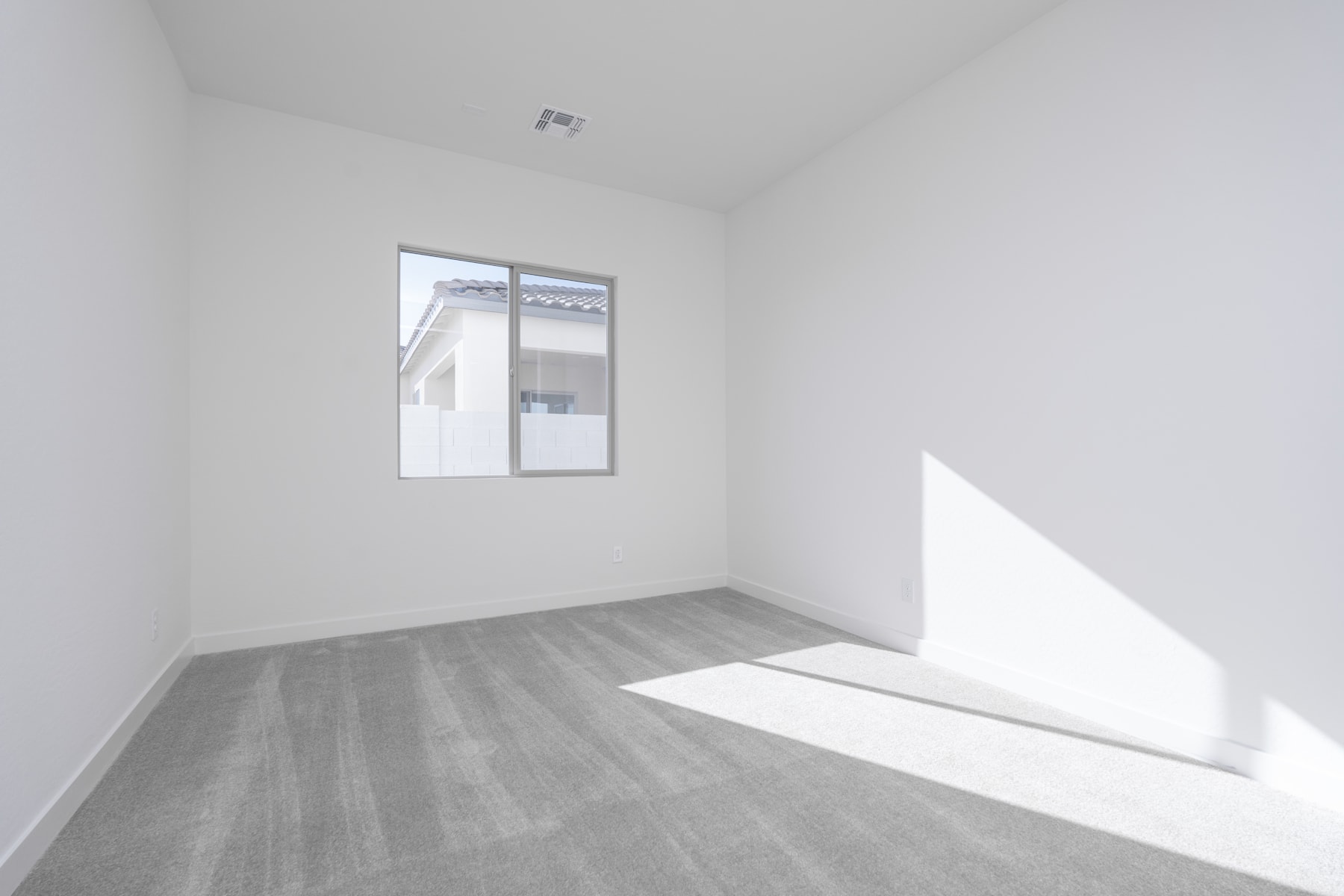 A minimalist, well-lit room with a large window, a plain white wall, and a gray hardwood floor.