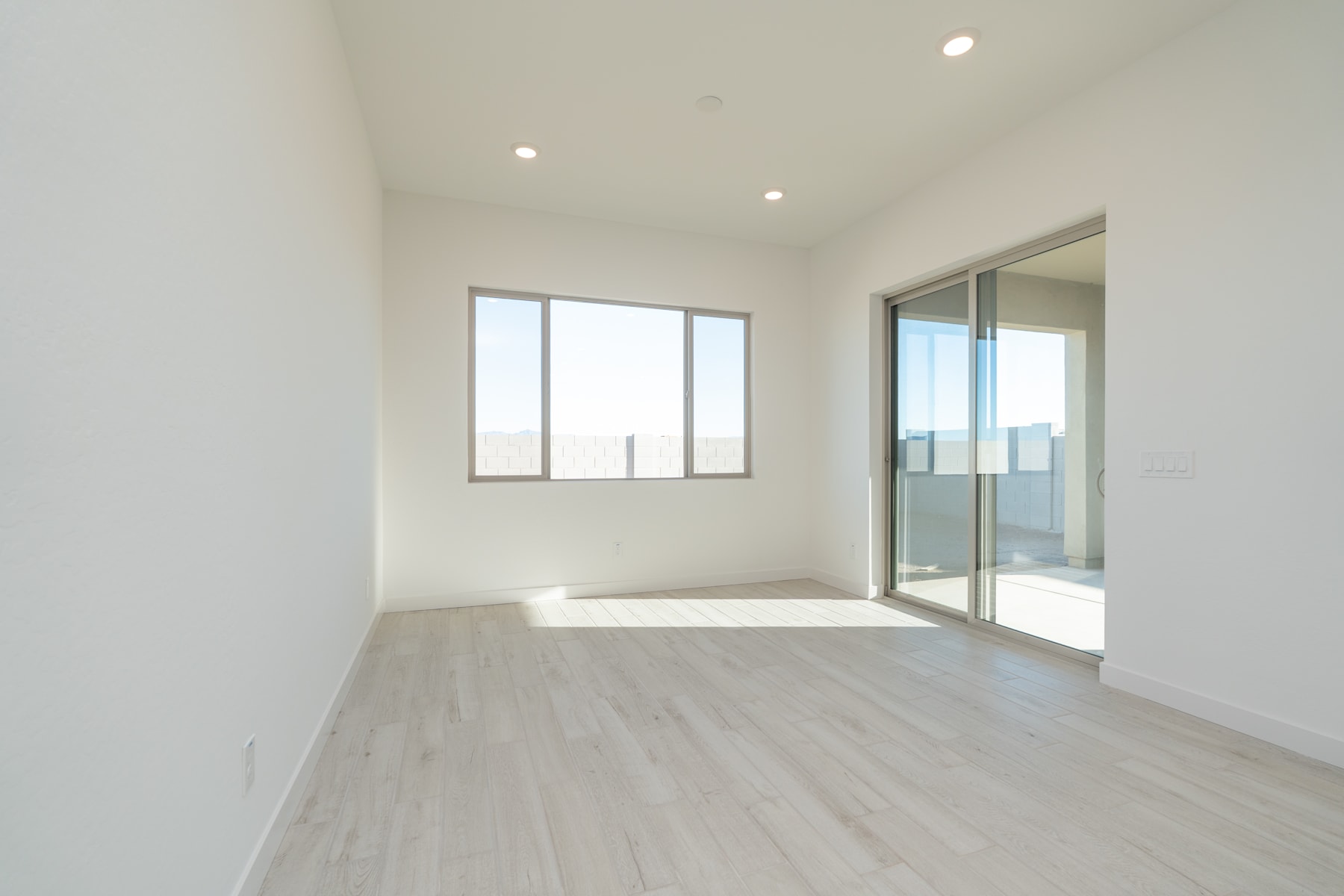 A bright, minimalist room with large windows, a sliding glass door, and a smooth, light-colored wooden floor.